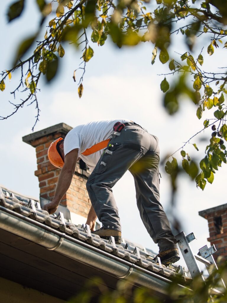 A worker in a helmet stands on a ladder fixing a roof near brick chimneys—contact us for a free roofing estimate Biloxi residents trust.