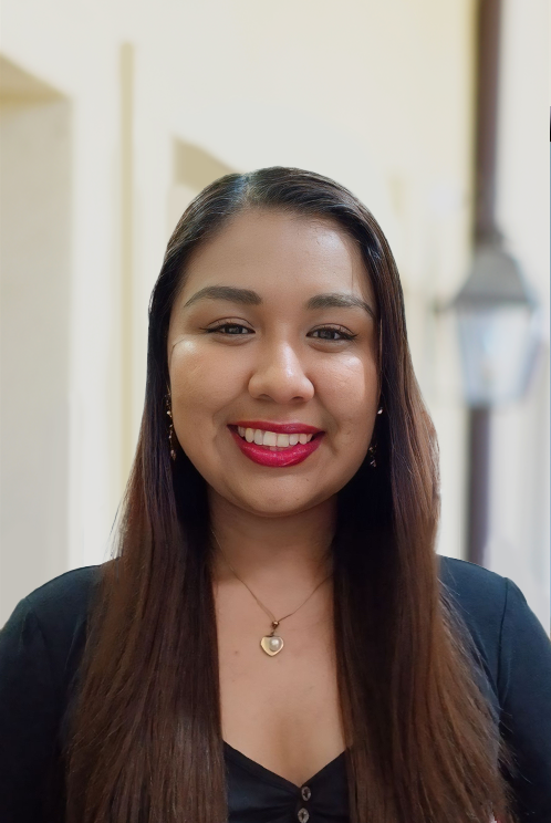 A woman with long brown hair, wearing a black top and a heart-shaped necklace, smiles indoors—ready to inspire you to join our team.