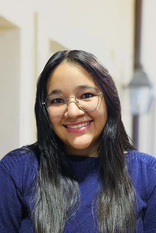 A woman with long dark hair and glasses smiles at the camera in a navy sweater—join our team and share this welcoming energy.