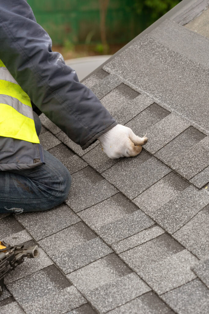 A person in gloves and a high-visibility vest repairs gray asphalt roof shingles—contact us for a free roofing estimate Biloxi.