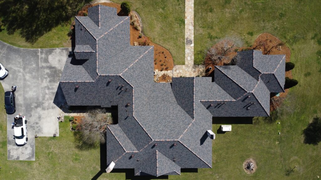 Aerial view of a large house with an Owens Corning roof deployment, featuring a dark gray roof, driveway, and several parked cars.